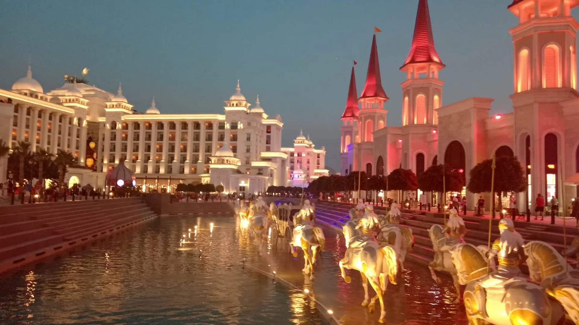 The Land of Legends at dusk: floodlit white towers with red spires beside a canal where gilded horses and riders stand in the water, and a lit hotel façade on the far bank