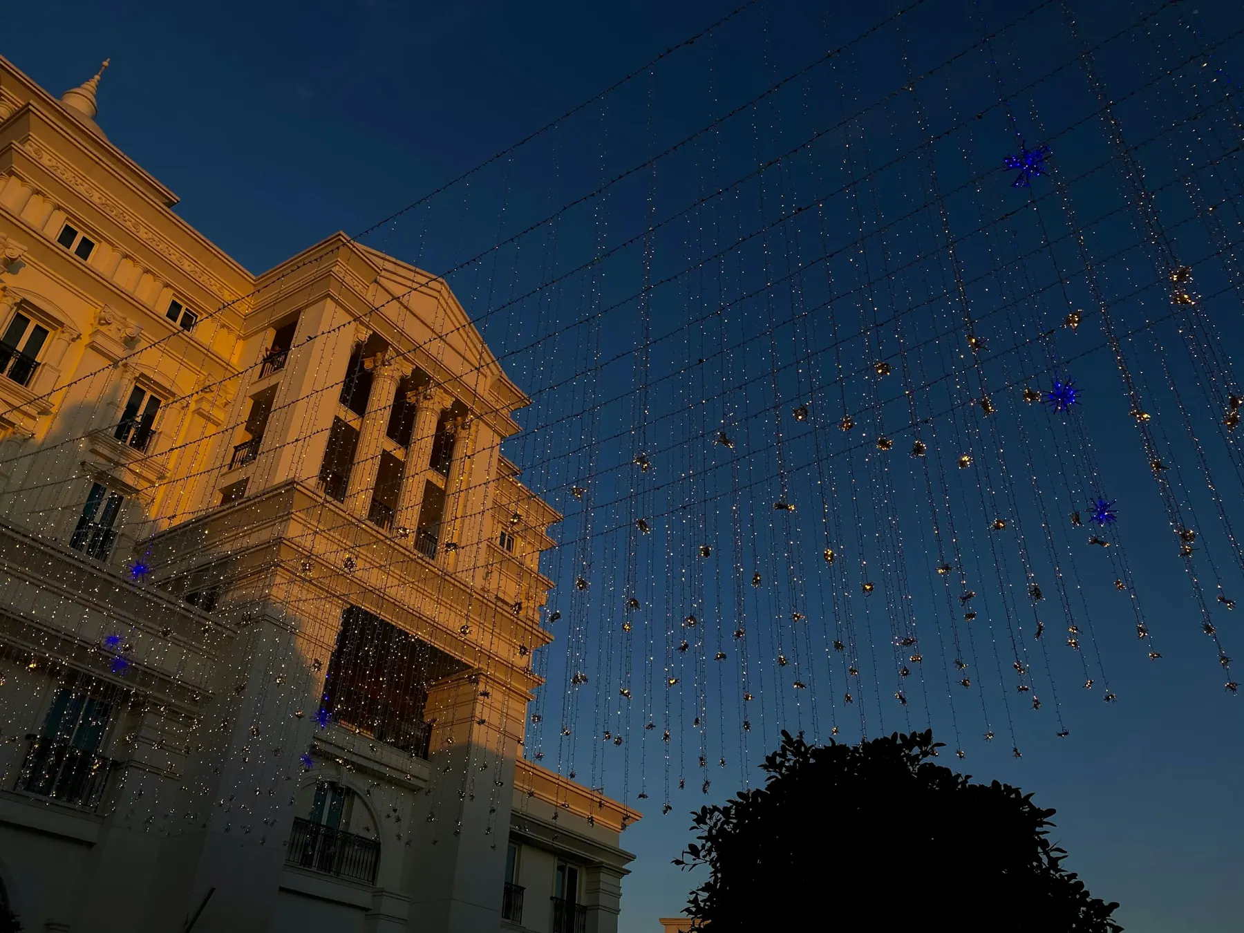 Strings of tiny lights hang across a dusk sky above a cream-coloured colonnaded building on the Shopping Avenue